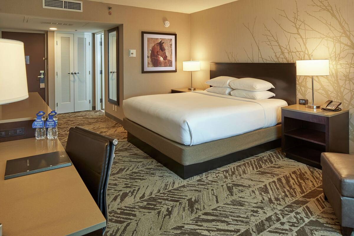 A hotel room with a bed, two bedside lamps, a desk with a chair, a water bottle, and a framed picture on the wall.
