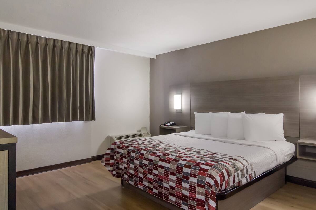 Ein Hotelzimmer mit einem großen Bett, einem Nachttisch, einem Sessel, einem Schreibtisch und einem Fenster mit Vorhängen.