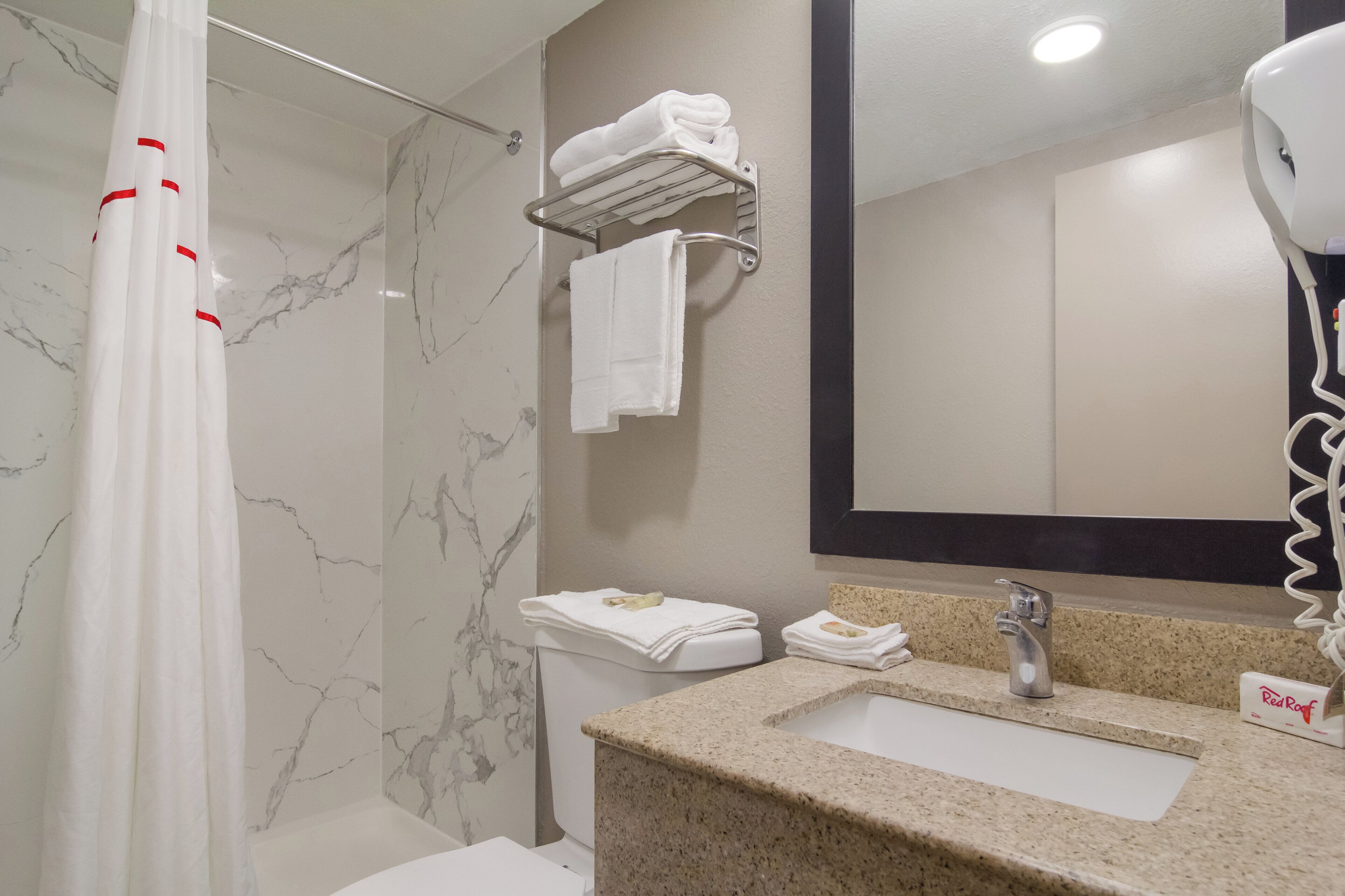 A bathroom with a marble wall, a shower curtain, a towel rack, a sink with a Red Roof logo, and a hairdryer.