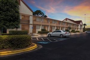 Un hotel con estacionamiento, un coche blanco aparcado y un letrero azul de estacionamiento para discapacitados.