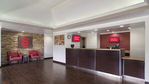 A hotel reception area with a stone wall, two red chairs, a reception desk, and a clock. - Red Roof Inn & Suites Brunswick I-95 (Brunswick)