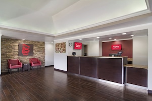 Zona de recepción de hotel con pared de piedra, dos sillas rojas, un mostrador de recepción y un reloj.
