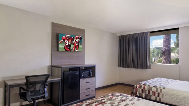 Ein Hotelzimmer mit Schreibtisch, einem an der Wand befestigten Fernseher, einem Bett mit Patchworkdecke und einem Fenster mit Vorhängen.