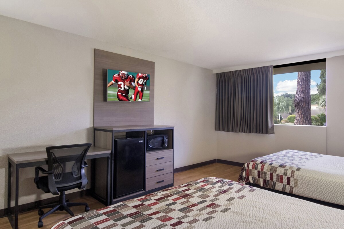 Habitación de hotel con escritorio, televisor montado en la pared, cama con colcha y ventana con cortinas.