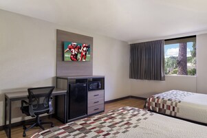 Habitación de hotel con escritorio, televisor montado en la pared, cama con colcha y ventana con cortinas.