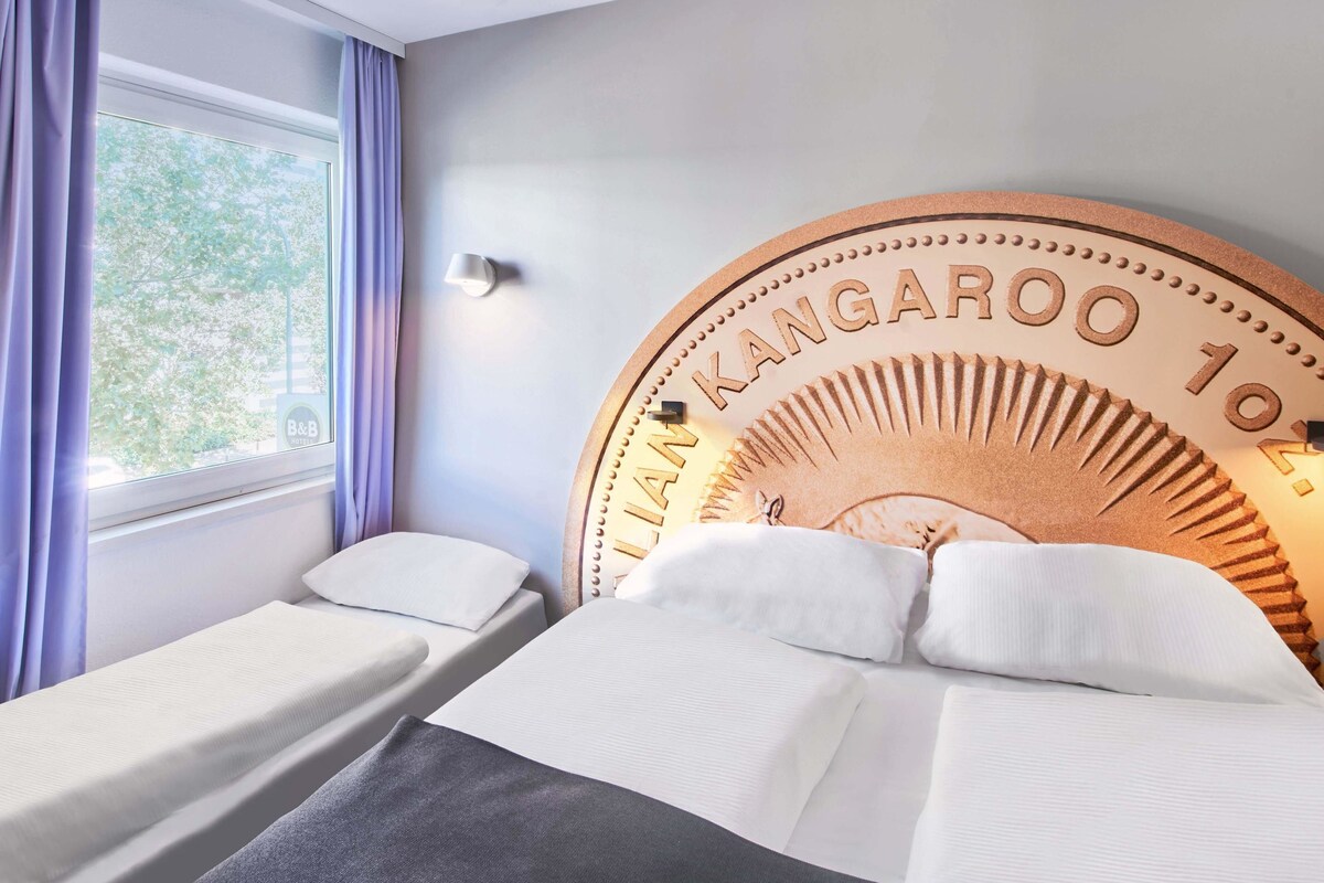 Una habitación de hotel con dos camas individuales, un cabecero de madera grande con la inscripción 'LANK KANGAROO 10' y una ventana con cortinas, además de una lámpara fijada a la pared.