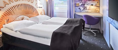 A hotel room with a bed, a desk, and a chair. The bed has a headboard with the text "ANGAROO". There is a window with curtains, and a poster on the wall.