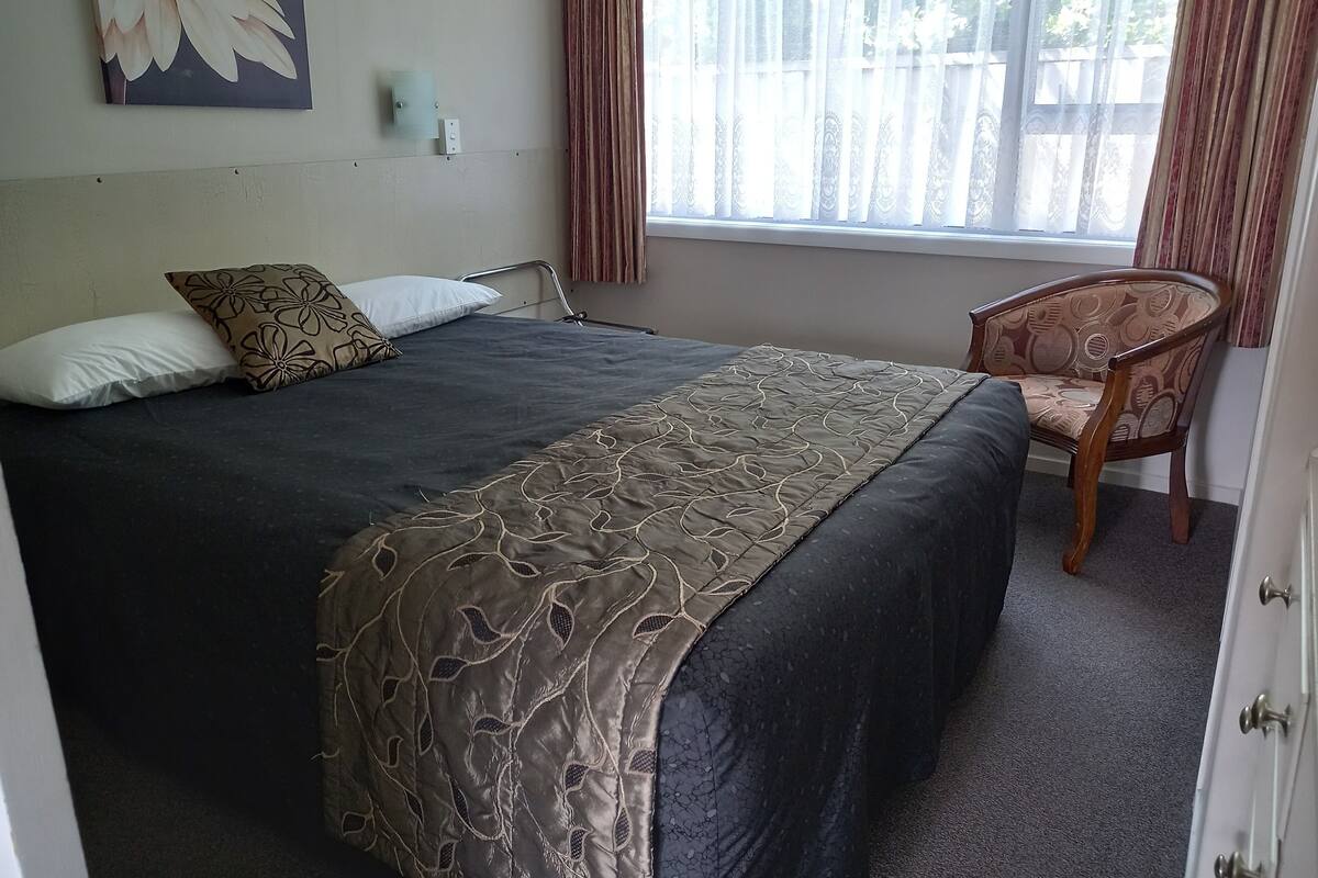 A neatly made bed with a dark bedspread and a decorative pillow, a wooden chair with a patterned cushion, and a window with sheer curtains.