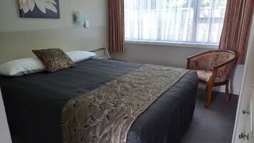 Un lit bien fait, recouvert d’une couvre-lit sombre et agrémenté d’un coussin décoratif, une chaise en bois avec un coussin à motifs et une fenêtre ornée de rideaux légers.