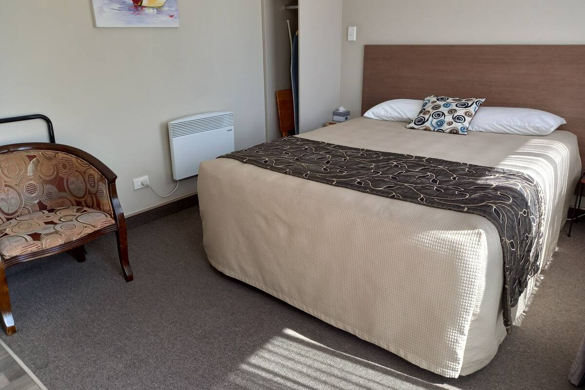 A hotel room with a bed, a chair, a heater, and a painting on the wall.