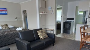 A modern living room with a black leather sofa, a dining area with wooden chairs, a kitchen with a stove, and a bedroom with a bed and a painting. - Comfort Inn Tayesta (Invercargill)
