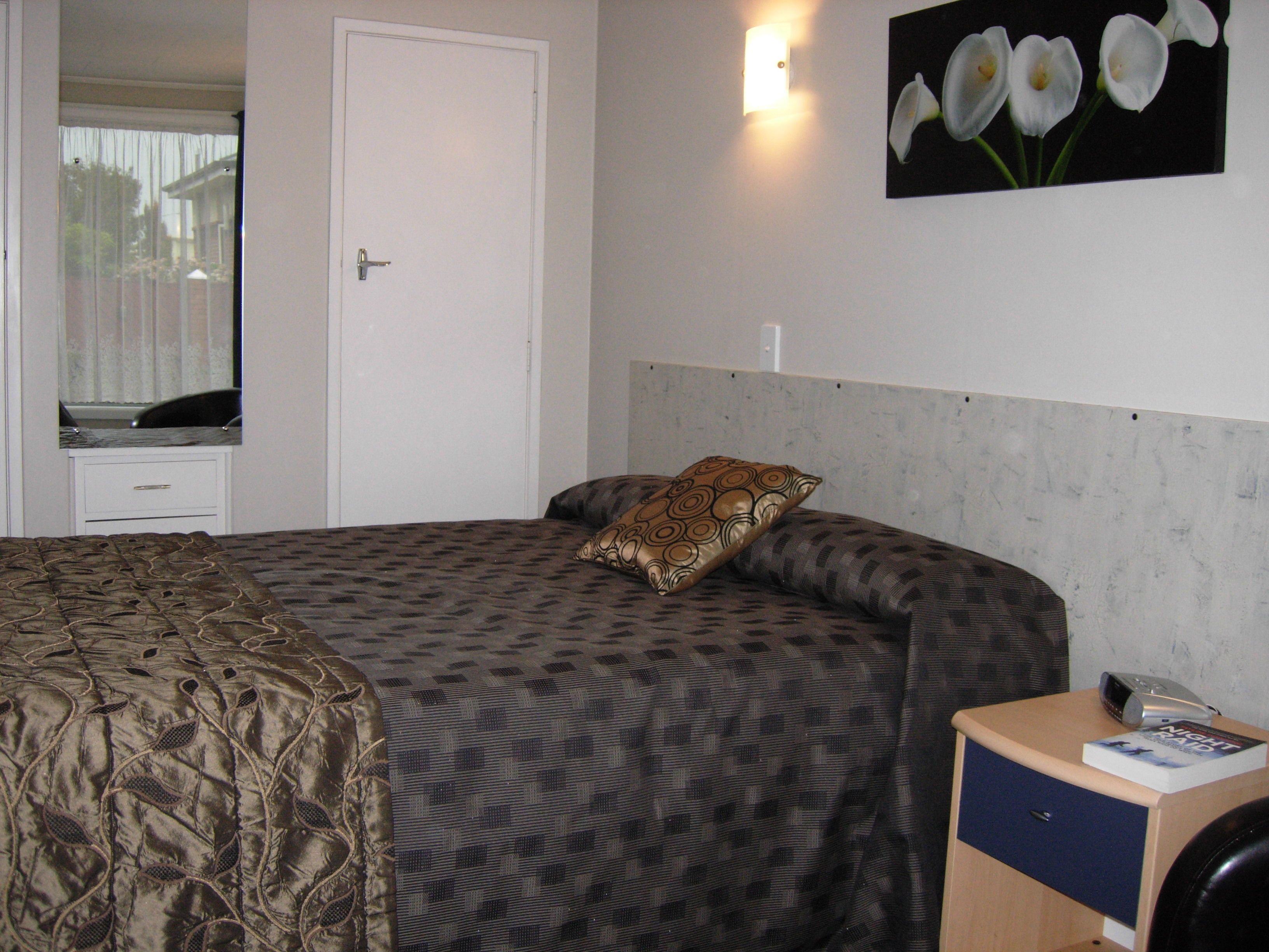 A bedroom with a bed, a nightstand, a drawer, a wall-mounted light, and a framed picture of flowers.