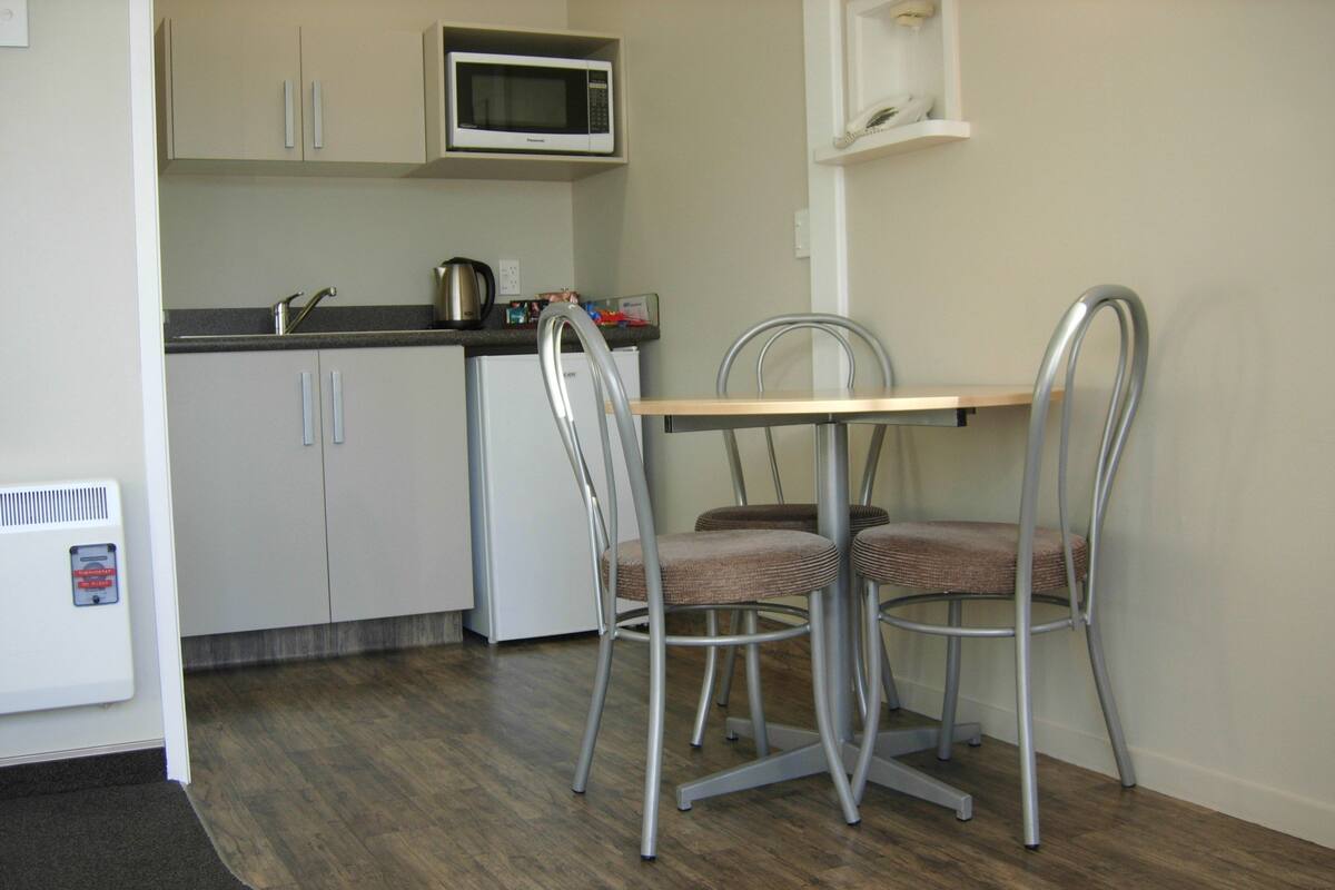 A compact kitchen with a microwave, kettle, and a small refrigerator. There are two metal chairs around a wooden table.