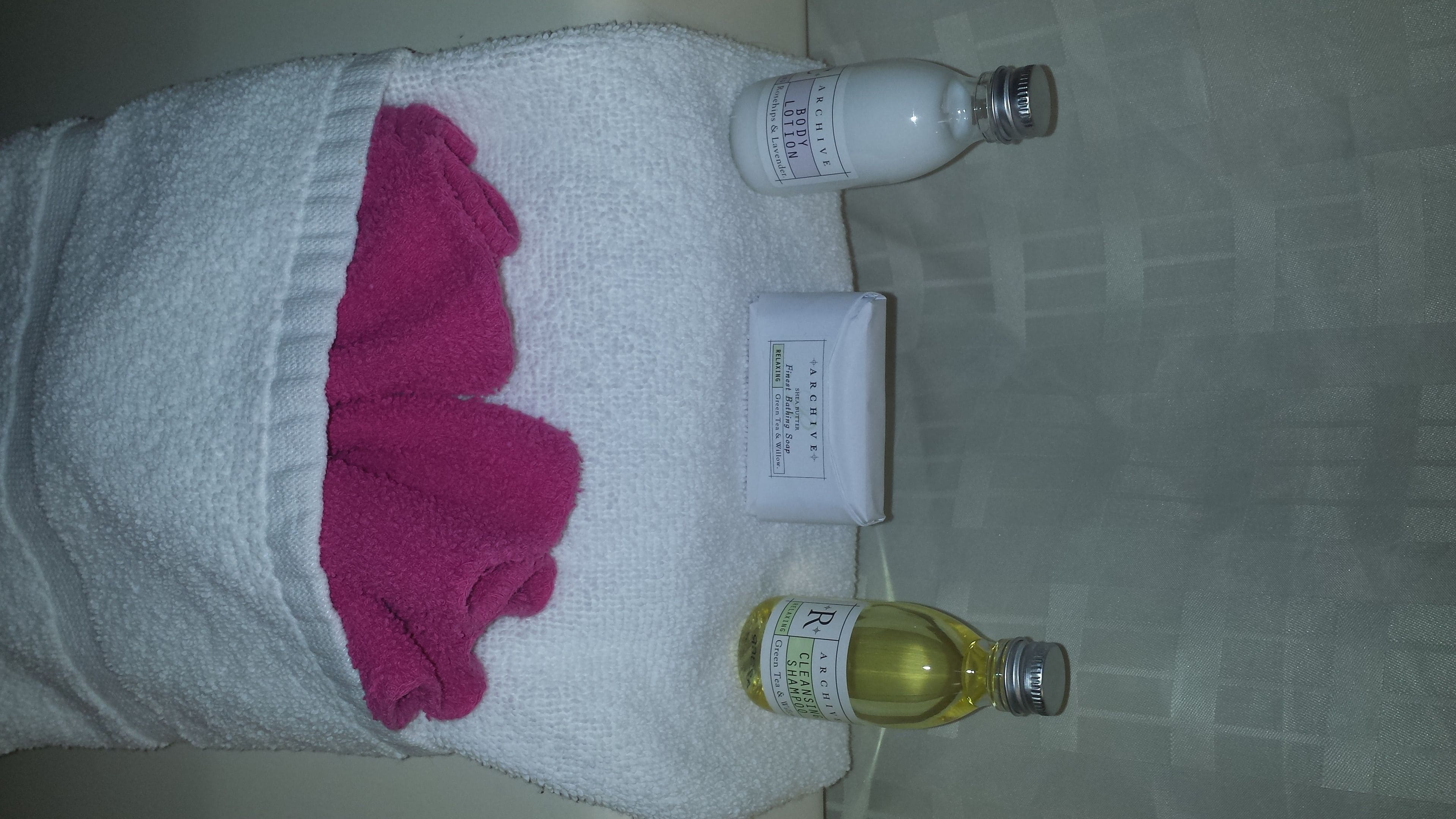 Une serviette blanche avec un mouchoir rose, un flacon de lotion pour le corps, un flacon de shampoing au thé vert et un flacon de gel douche, le tout posé sur un sol carrelé.
