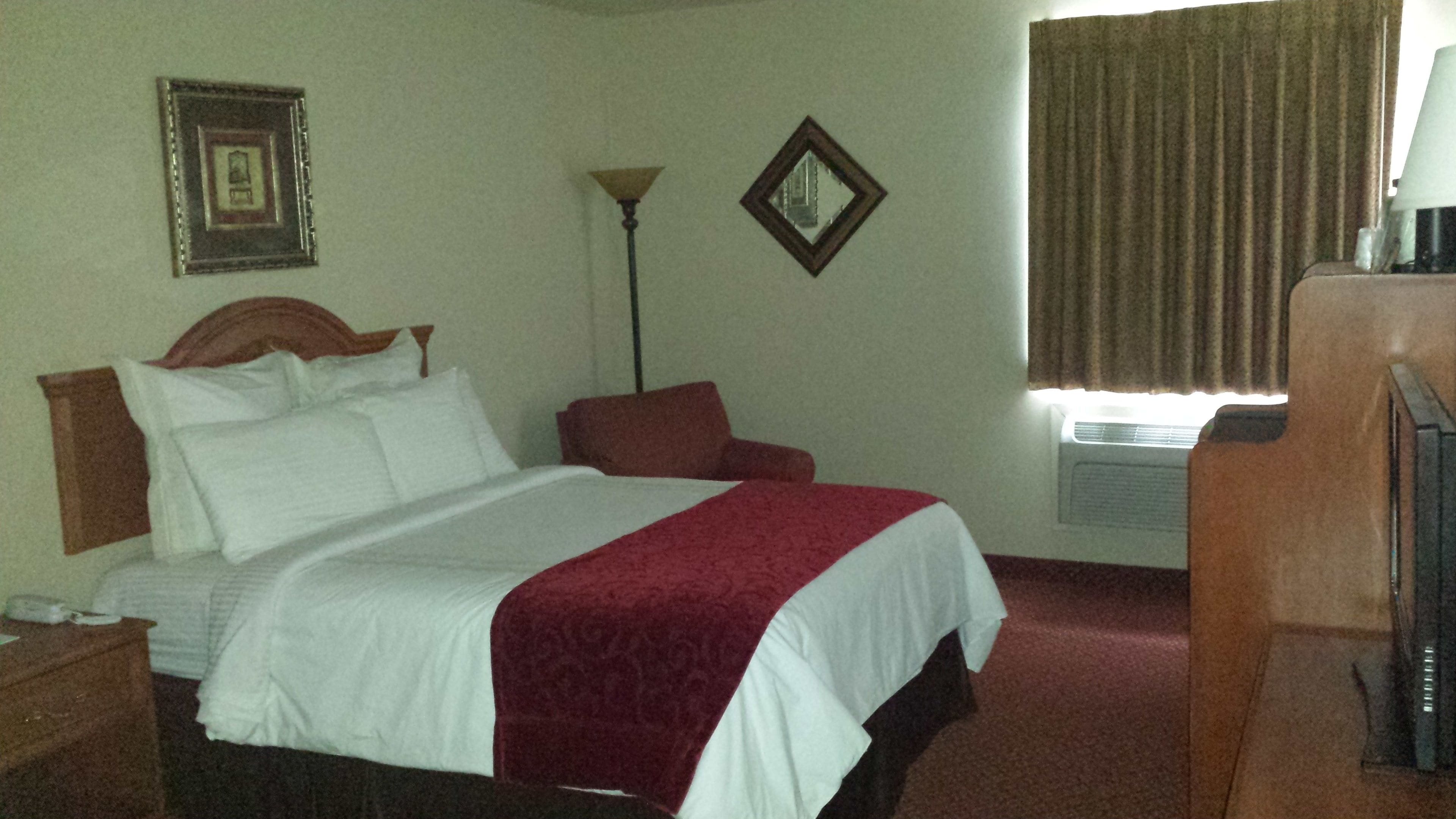 A hotel room with a bed, a chair, a lamp, a picture frame, and a window with curtains.