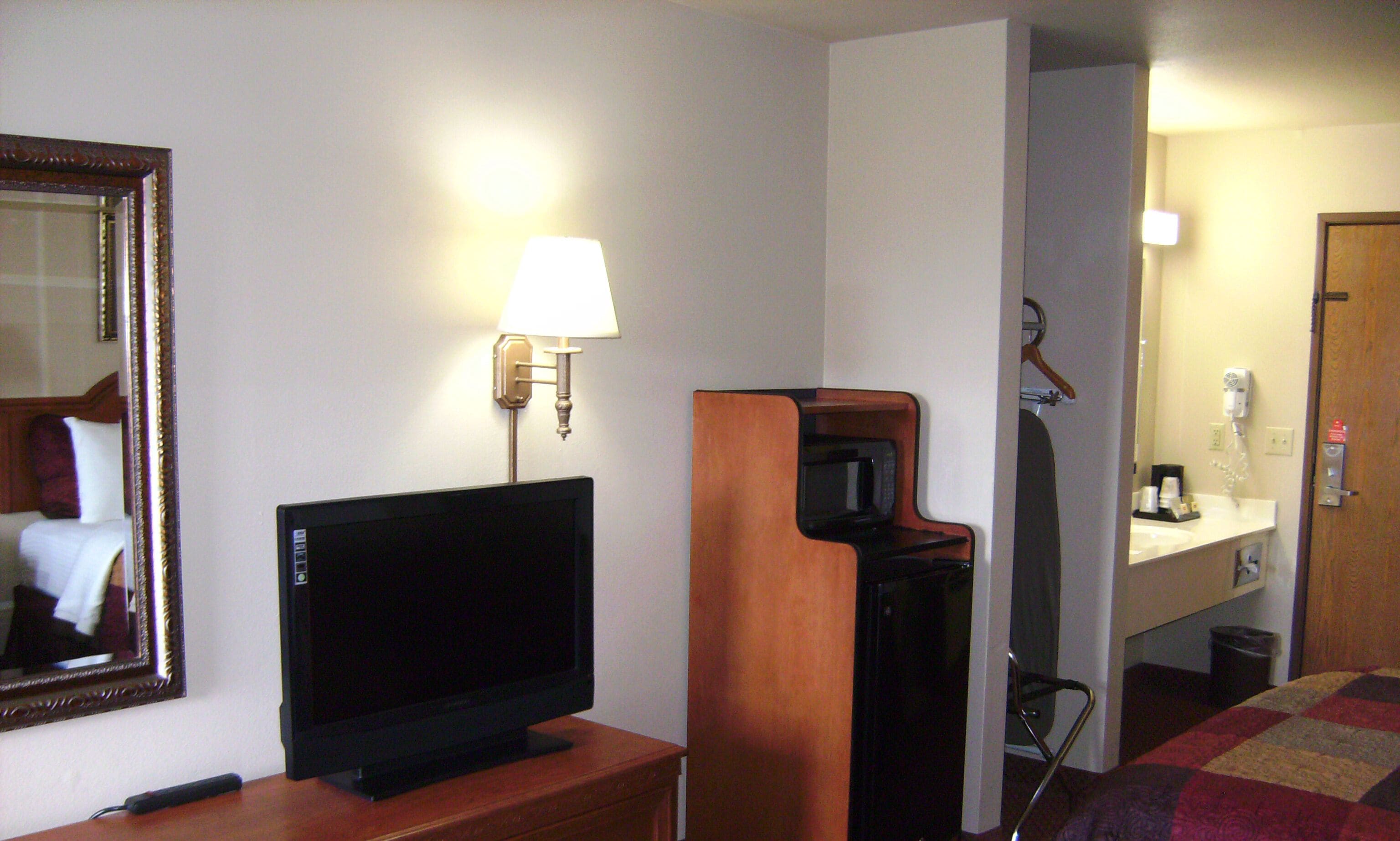 Ein Hotelzimmer mit Fernseher auf einem Holzständer, einer Wandlampe, einer Mikrowelle und einem kleinen Schreibtisch mit Spiegel.
