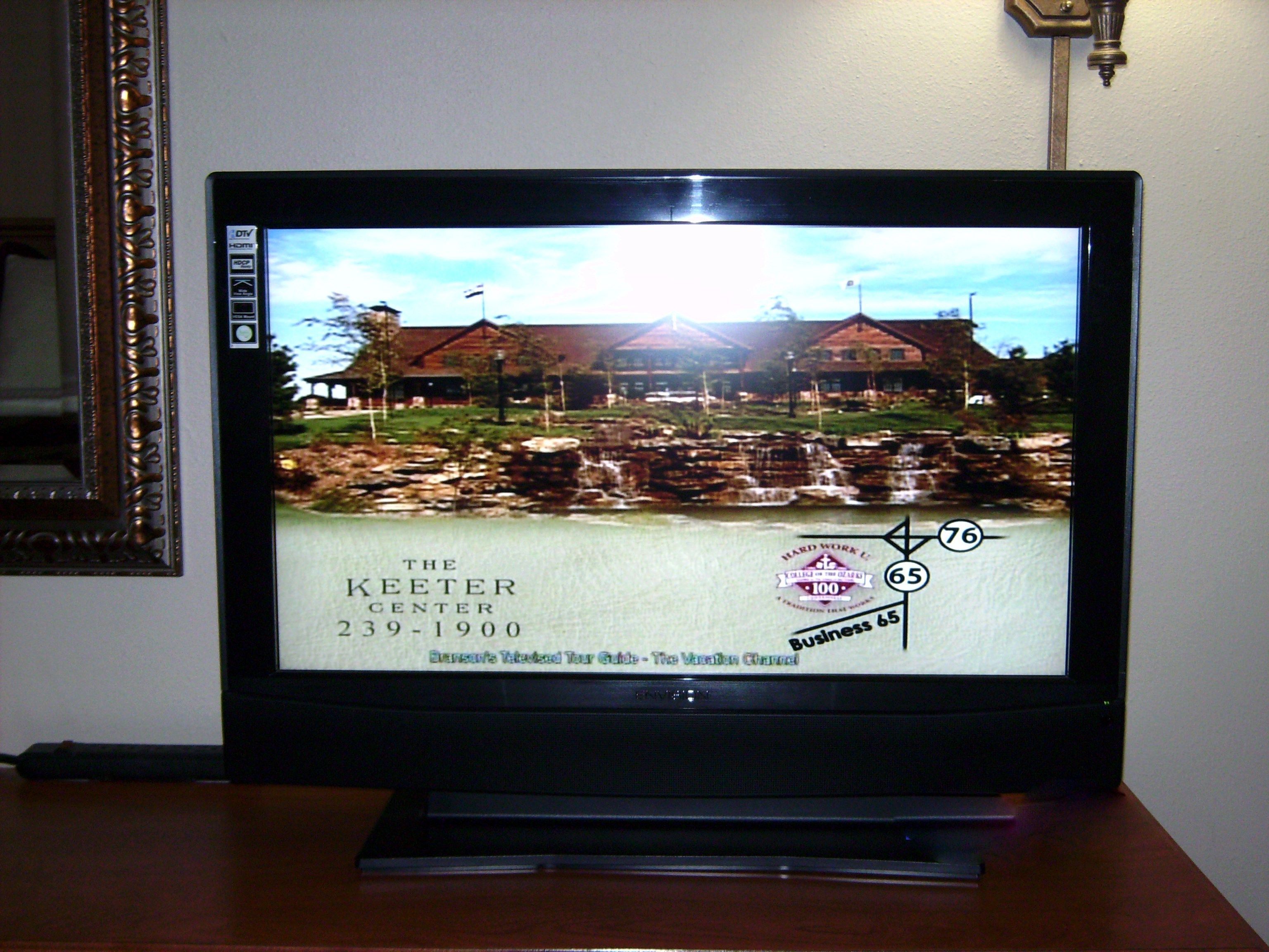 A television displaying a scenic view of a building with a sign reading "The Keeter Center 239-1900".