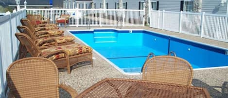 A swimming pool with wicker chairs and a table, surrounded by a white fence, with a building in the background.
