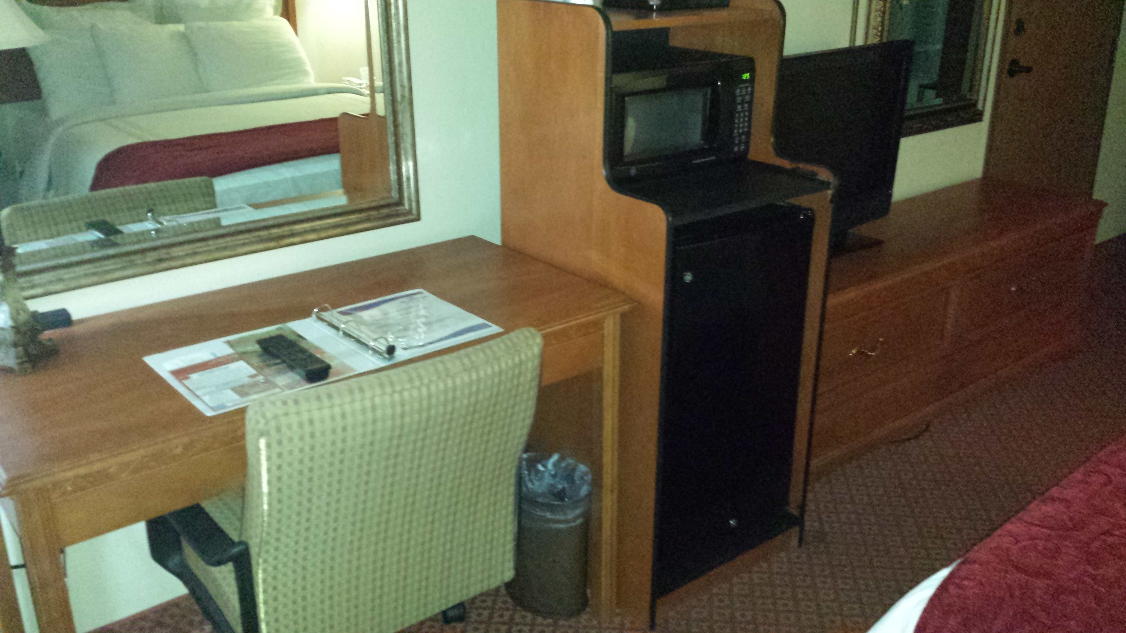 A compact hotel room with a wooden desk, a small black TV, a microwave, and a trash bin.