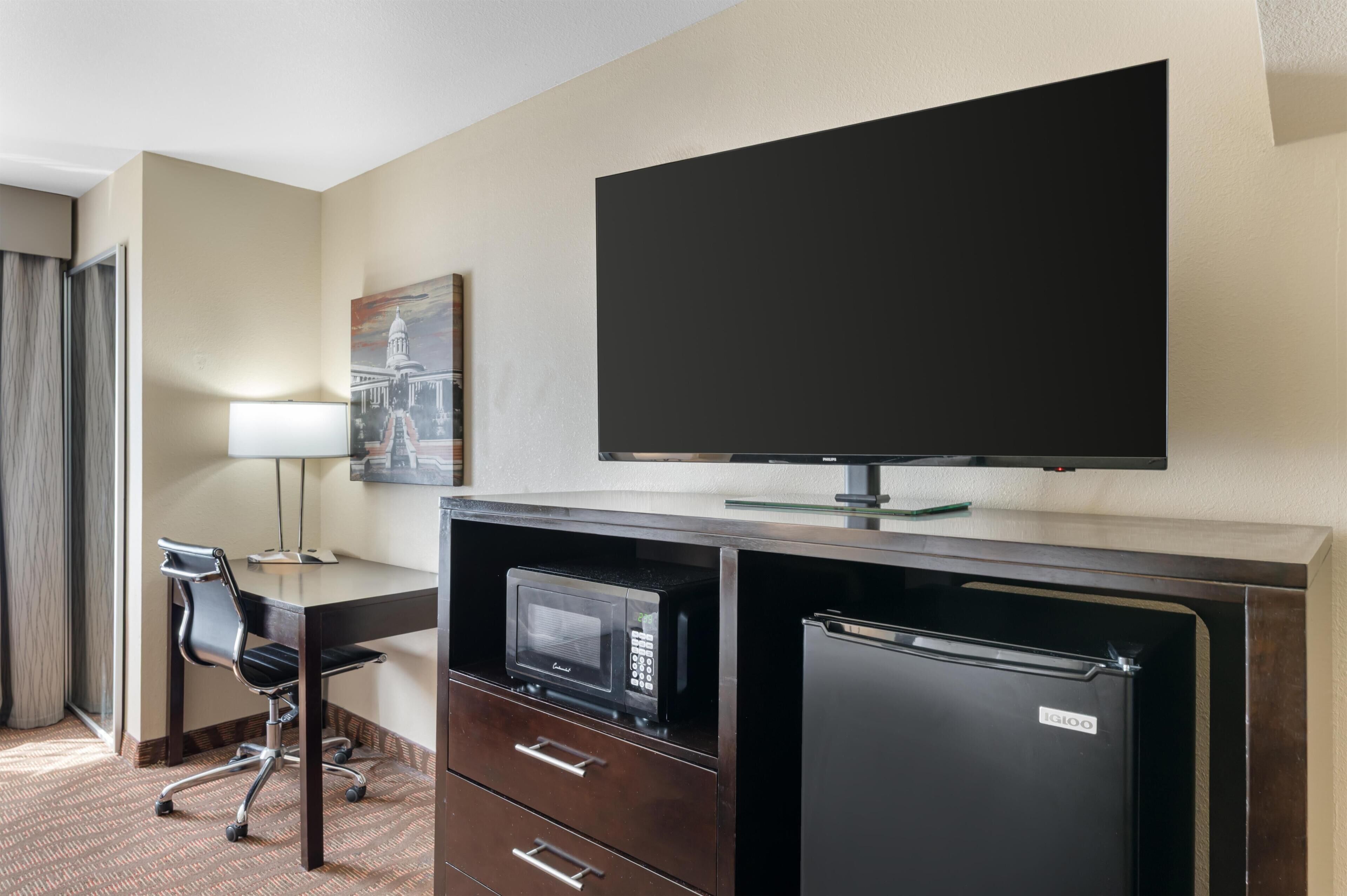 A hotel room with a desk, a flat-screen TV, a microwave, a refrigerator, a chair, and a painting on the wall.