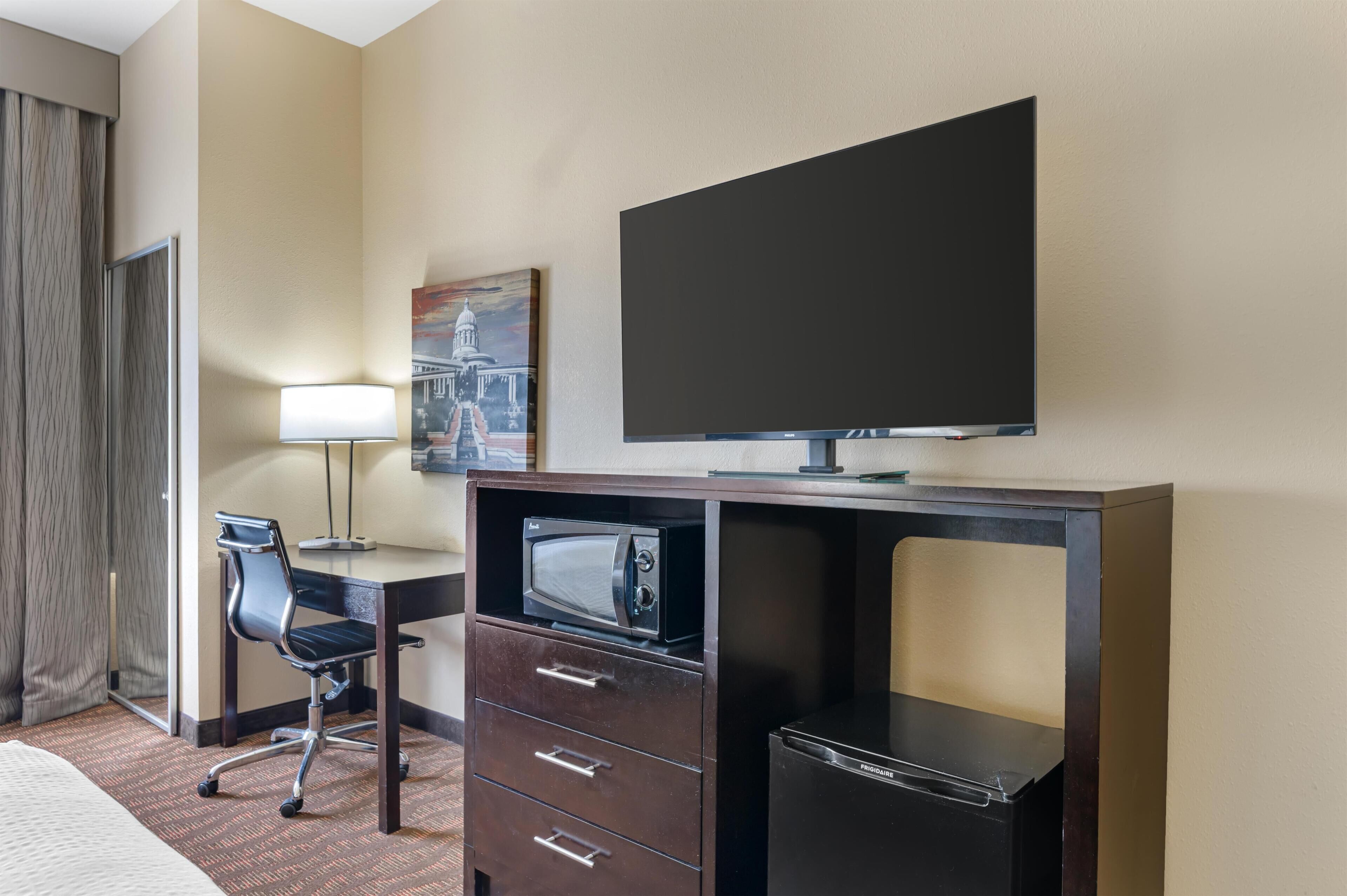 A hotel room with a bed, a desk with a computer, a microwave, a refrigerator, a TV, a chair, and a painting on the wall.