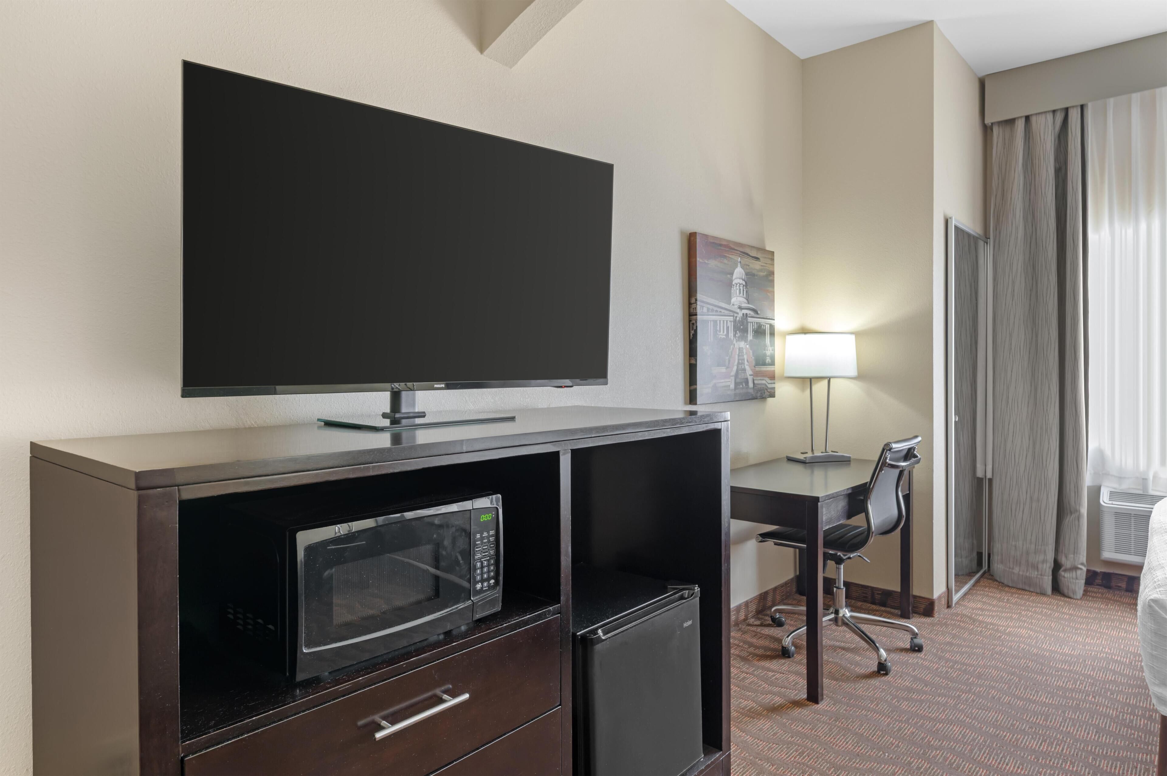 Habitación de hotel con televisión de pantalla plana, microondas, refrigerador, escritorio con silla, lámpara y una ventana grande con cortinas.
