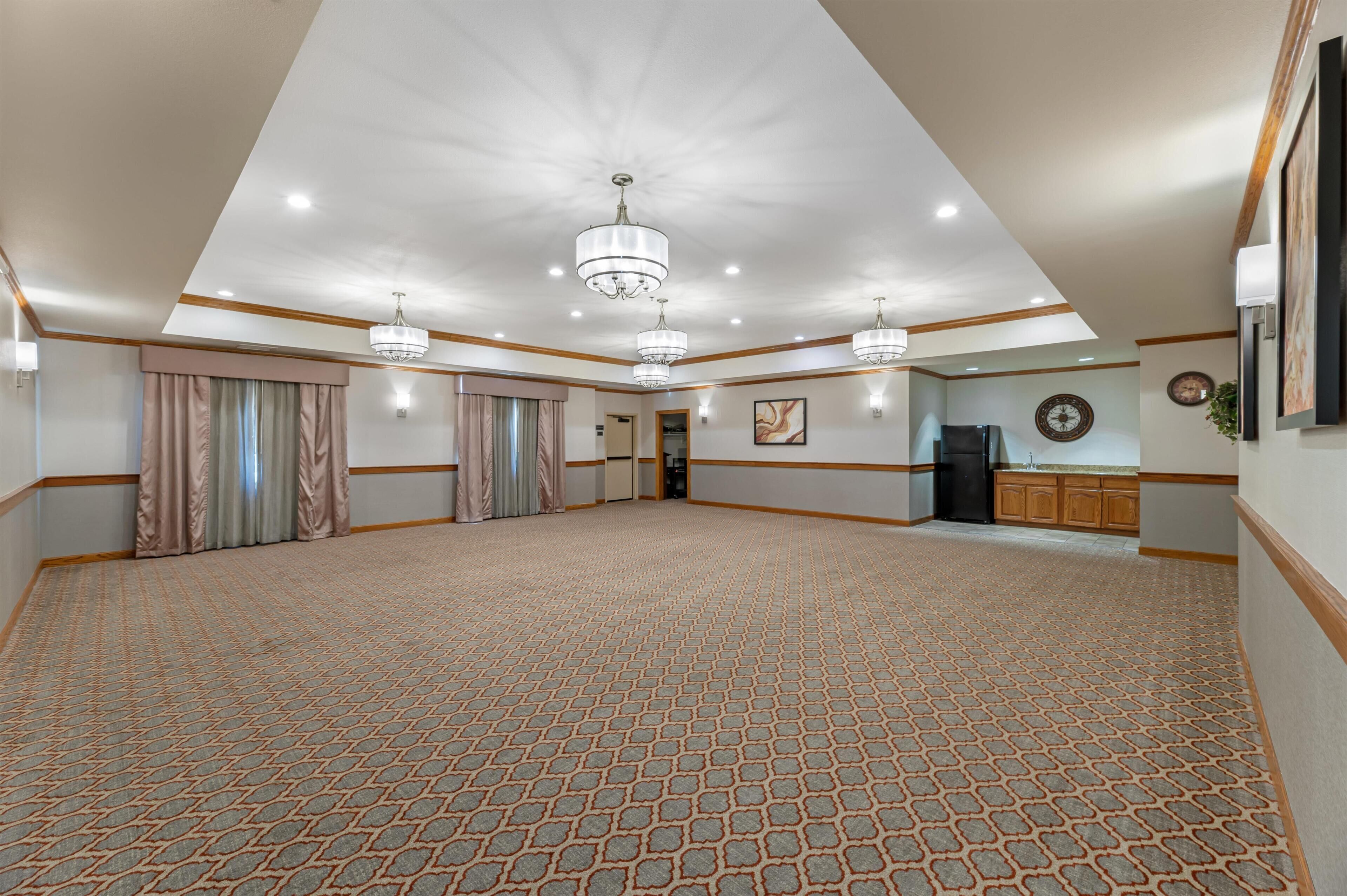 A spacious room with patterned carpet, chandeliers, a kitchen area, and a clock on the wall.
