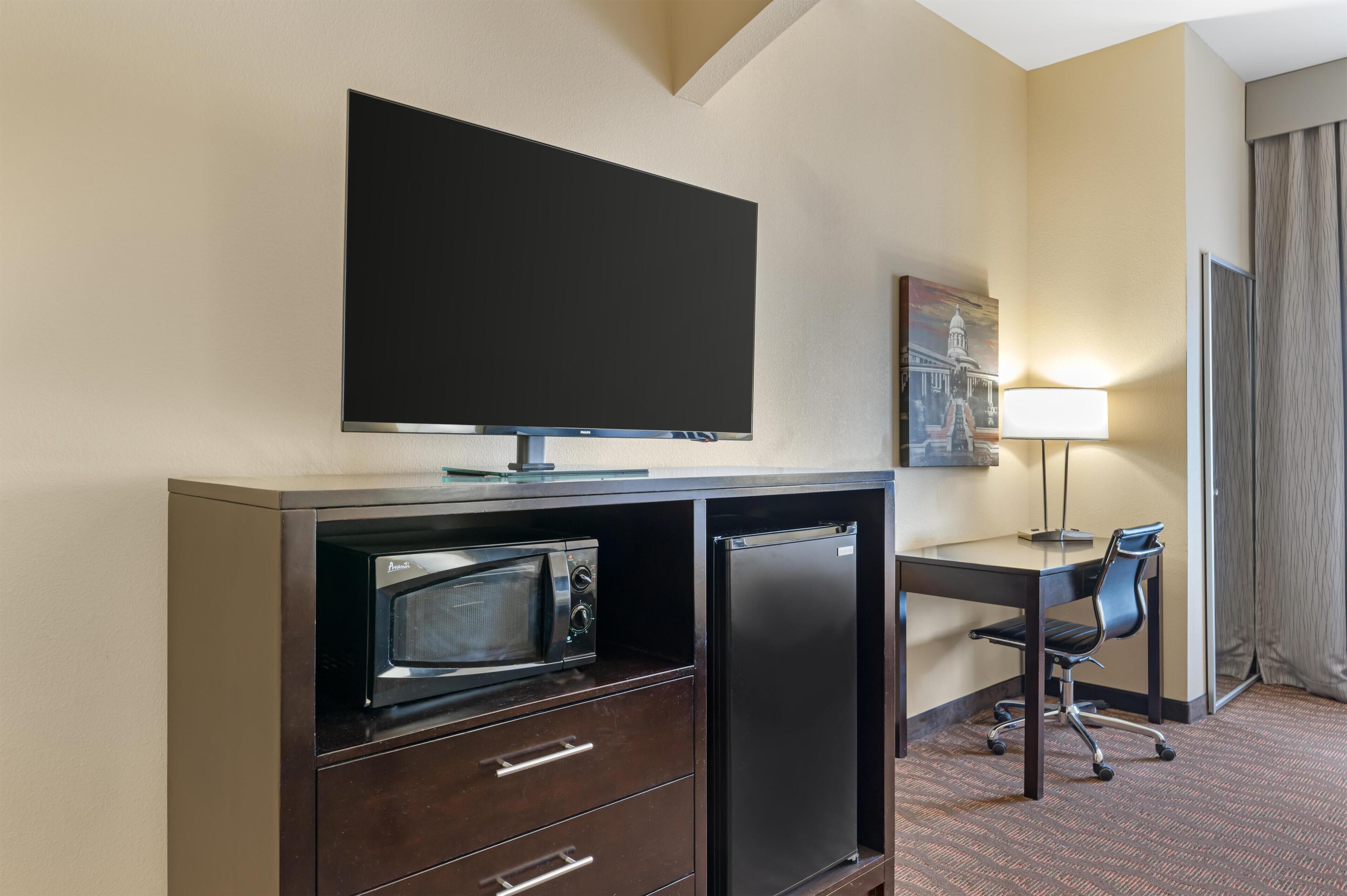 A hotel room with a flat-screen TV, a microwave, a refrigerator, a desk with a chair, and a lamp.