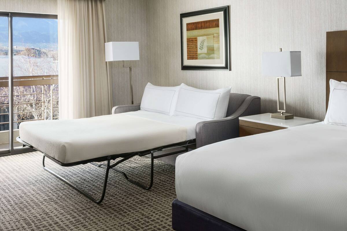 A hotel room with a bed, a bedside table, a lamp, a window with curtains, and a framed picture on the wall.
