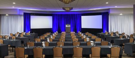 Un salón de conferencias con varias mesas y sillas, pantallas grandes y un escenario con un atril.