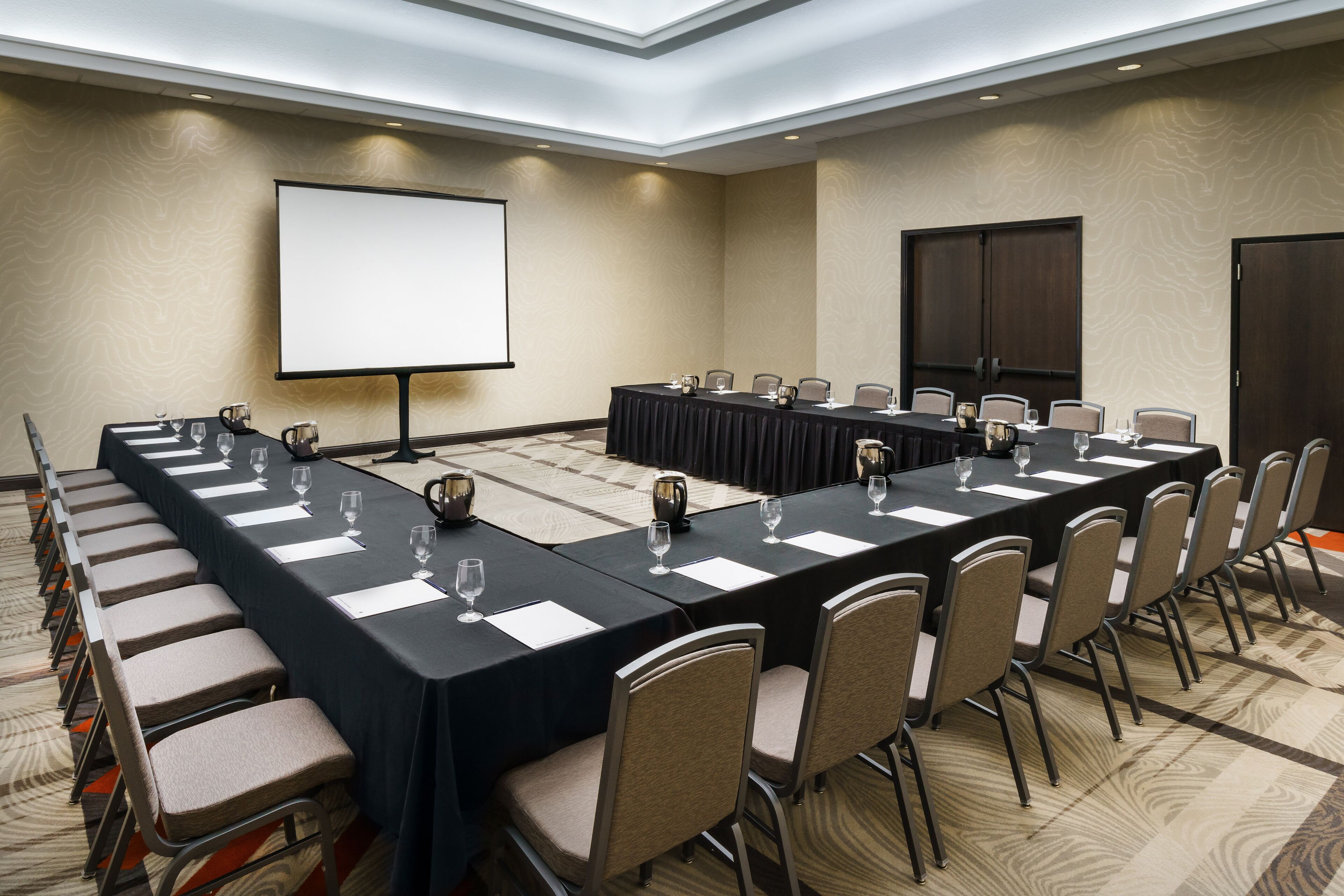 A conference room with a long table, chairs, a projector screen, and a carpeted floor.