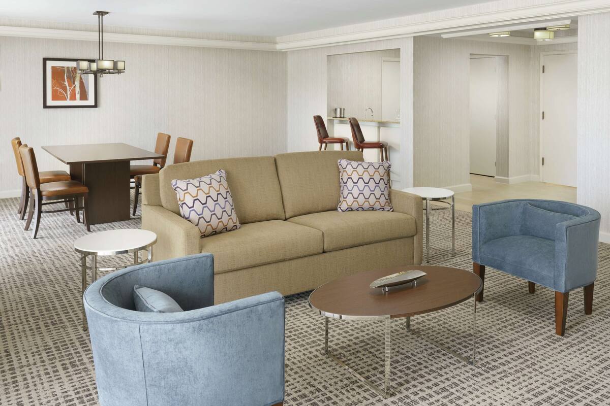 A modern hotel room with a beige sofa, two armchairs, a small round table, a dining area with a table and chairs, and a hallway with doors.