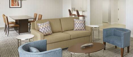 A modern hotel room with a beige sofa, two armchairs, a small round table, a dining area with a table and chairs, and a hallway with doors.