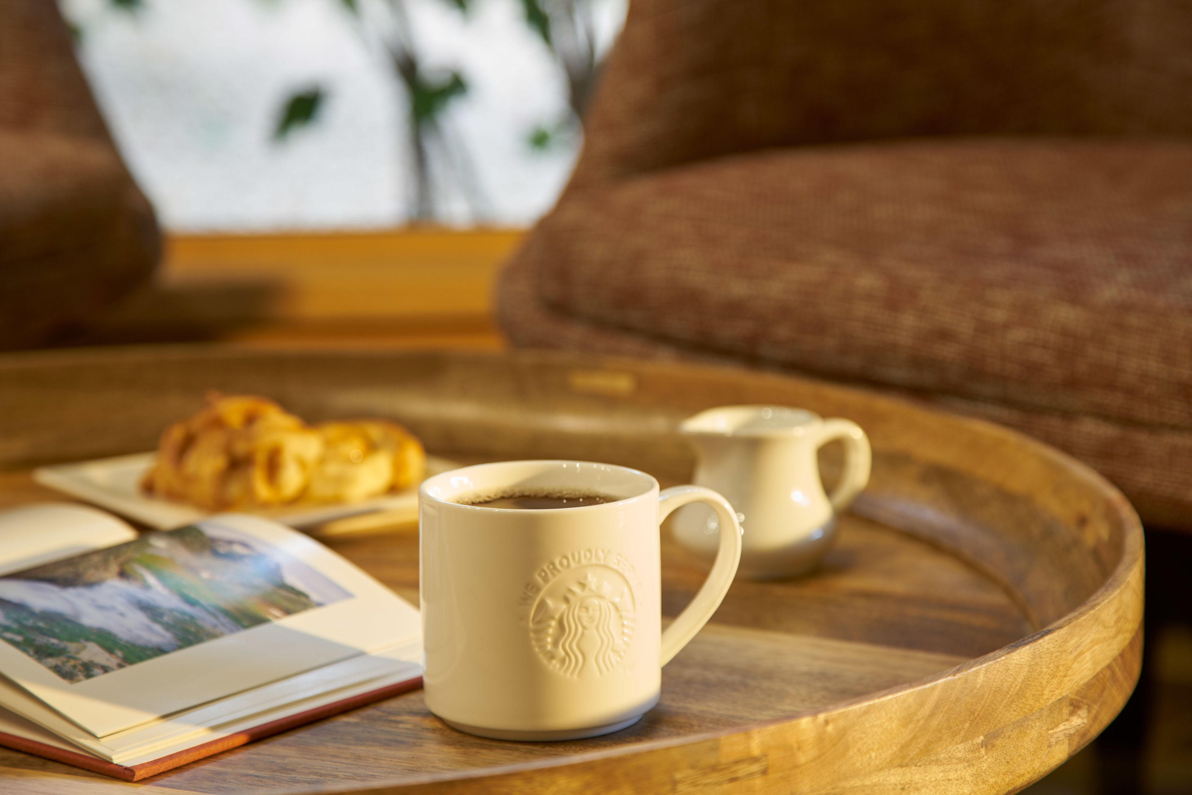 A wooden tray with a coffee mug and a croissant on a table, with a couch in the background.