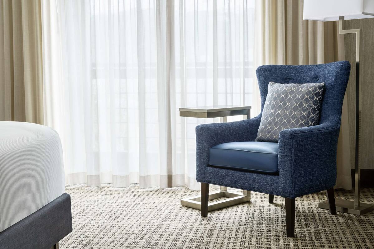 A hotel room with a blue armchair, a small side table, and a bed with a patterned bedspread.