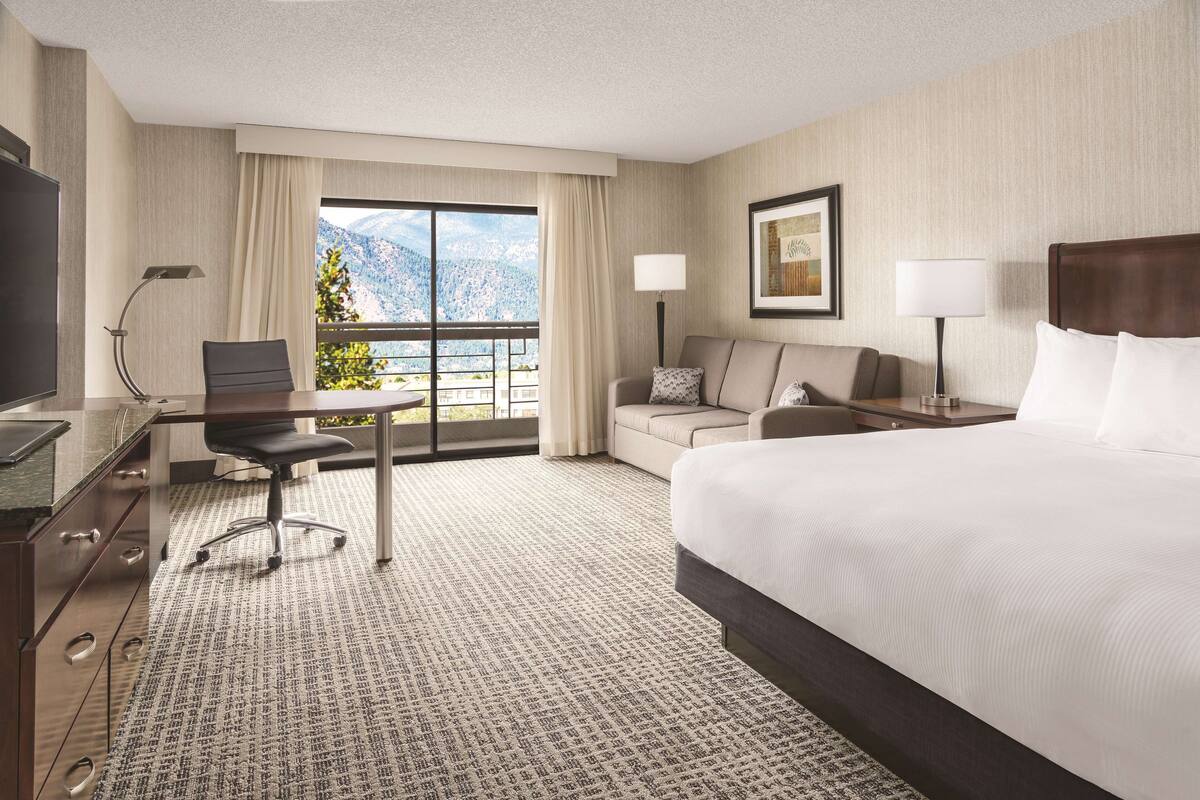 A hotel room with a large bed, a desk with a chair, a sofa, a TV, and a window with a mountain view.