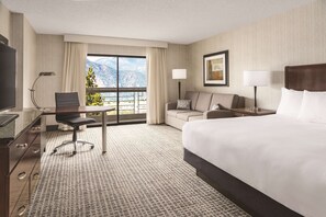 A hotel room with a large bed, a desk with a chair, a sofa, a TV, and a window with a mountain view.