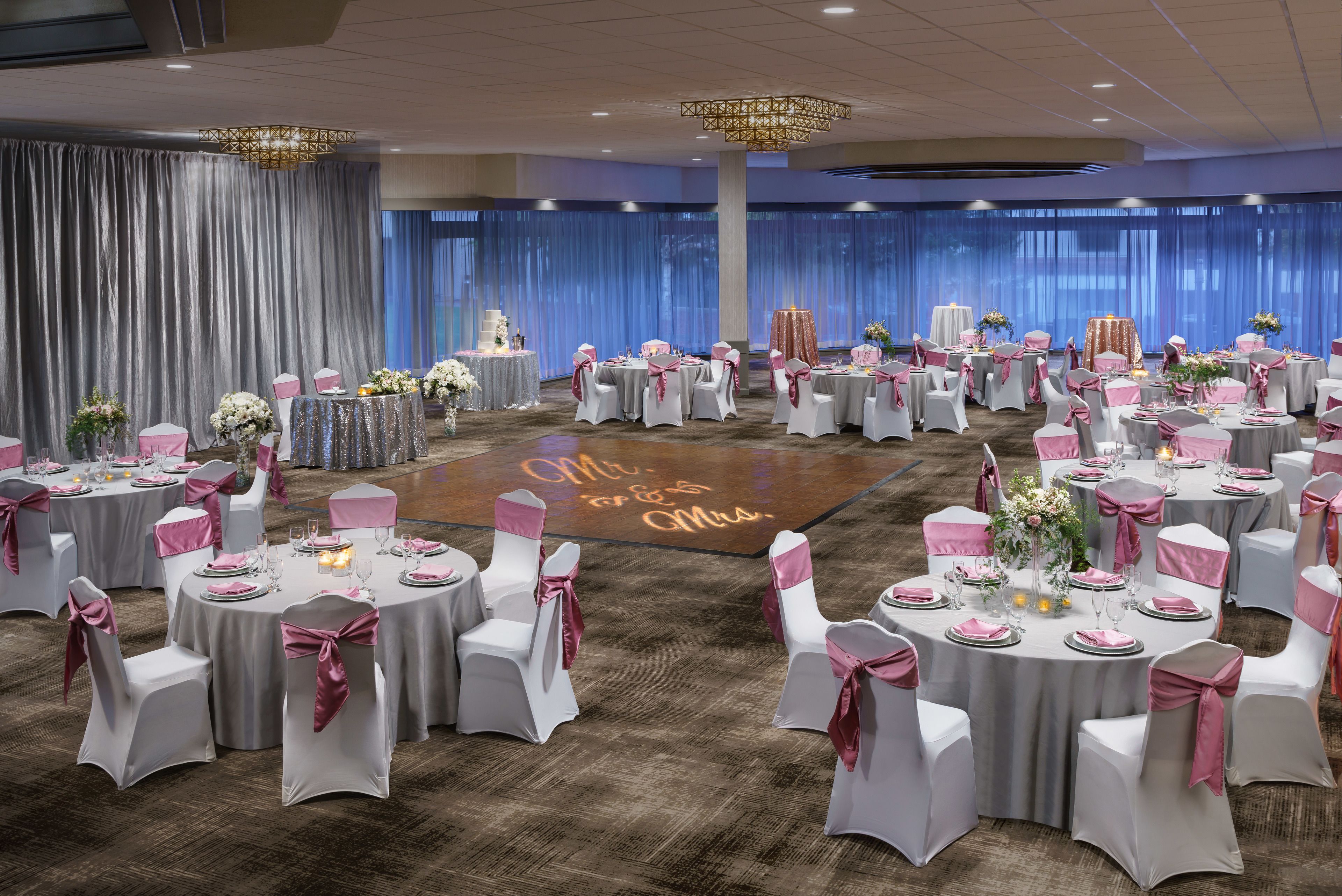 Ein Festsaal mit runden Tischen, weißen Stühlen mit rosa Bändern und einer Tanzfläche mit einem Neon-Schild.