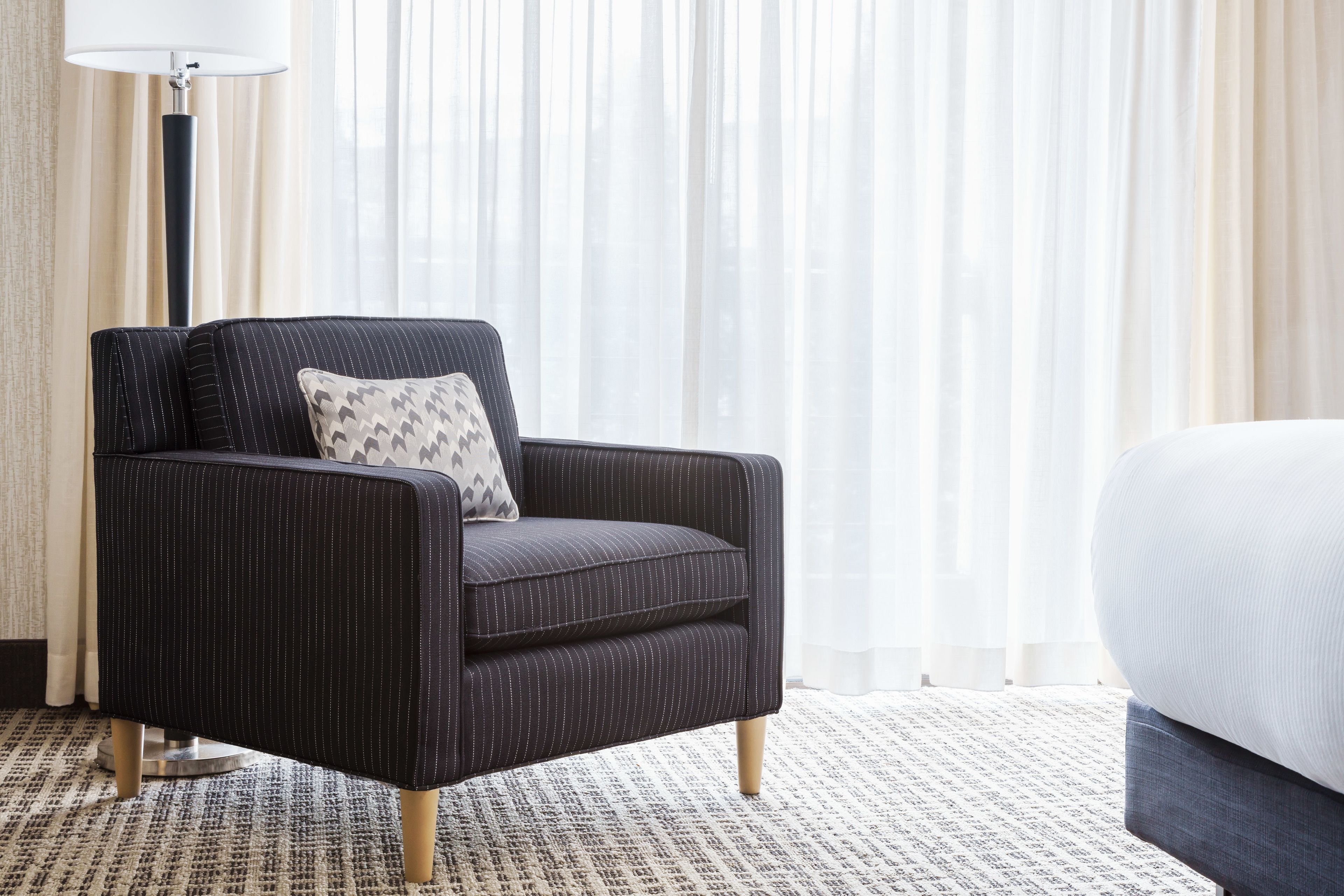 A dark upholstered armchair with a patterned pillow, a floor lamp, and a bed with a white headboard in a hotel room with a patterned carpet and sheer curtains.