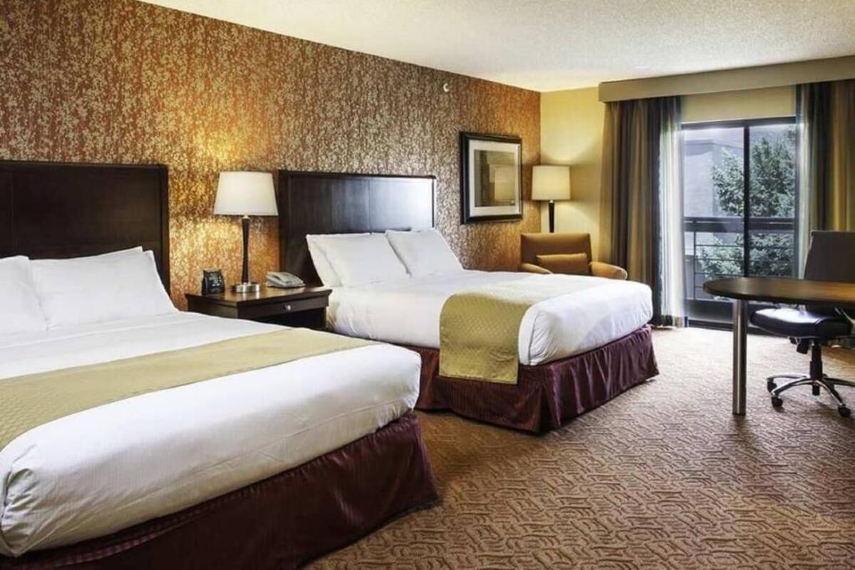 A hotel room with two beds, a desk, a chair, a lamp, and a window with a view.