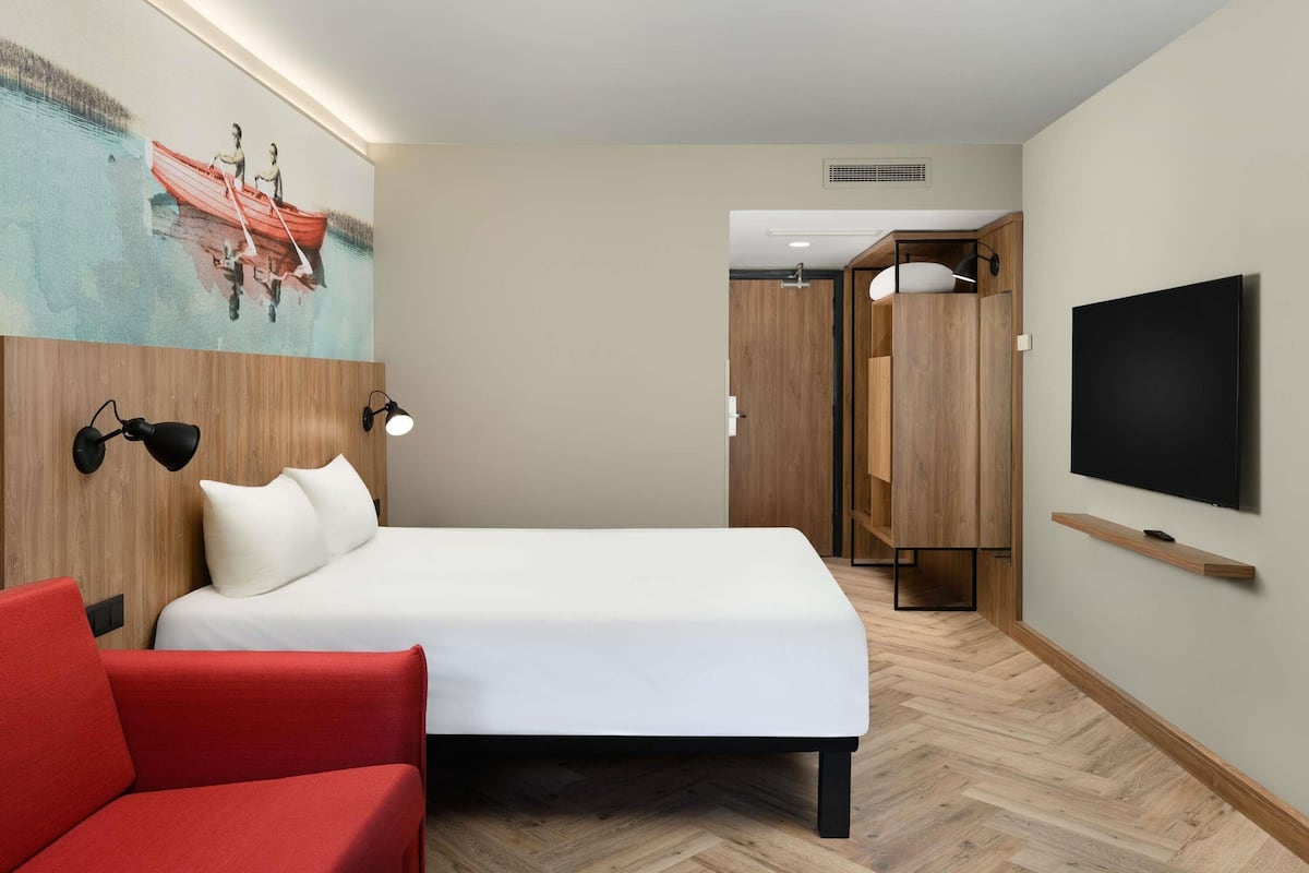 A hotel room with a bed, a red chair, a TV, and a painting on the wall.