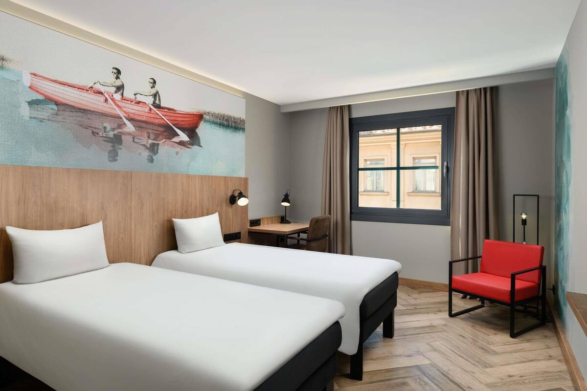 Hotellihuone, jossa on kaksi sänkyä, työpöytä, tuoli, taulu ja ikkuna, jossa on verhot.