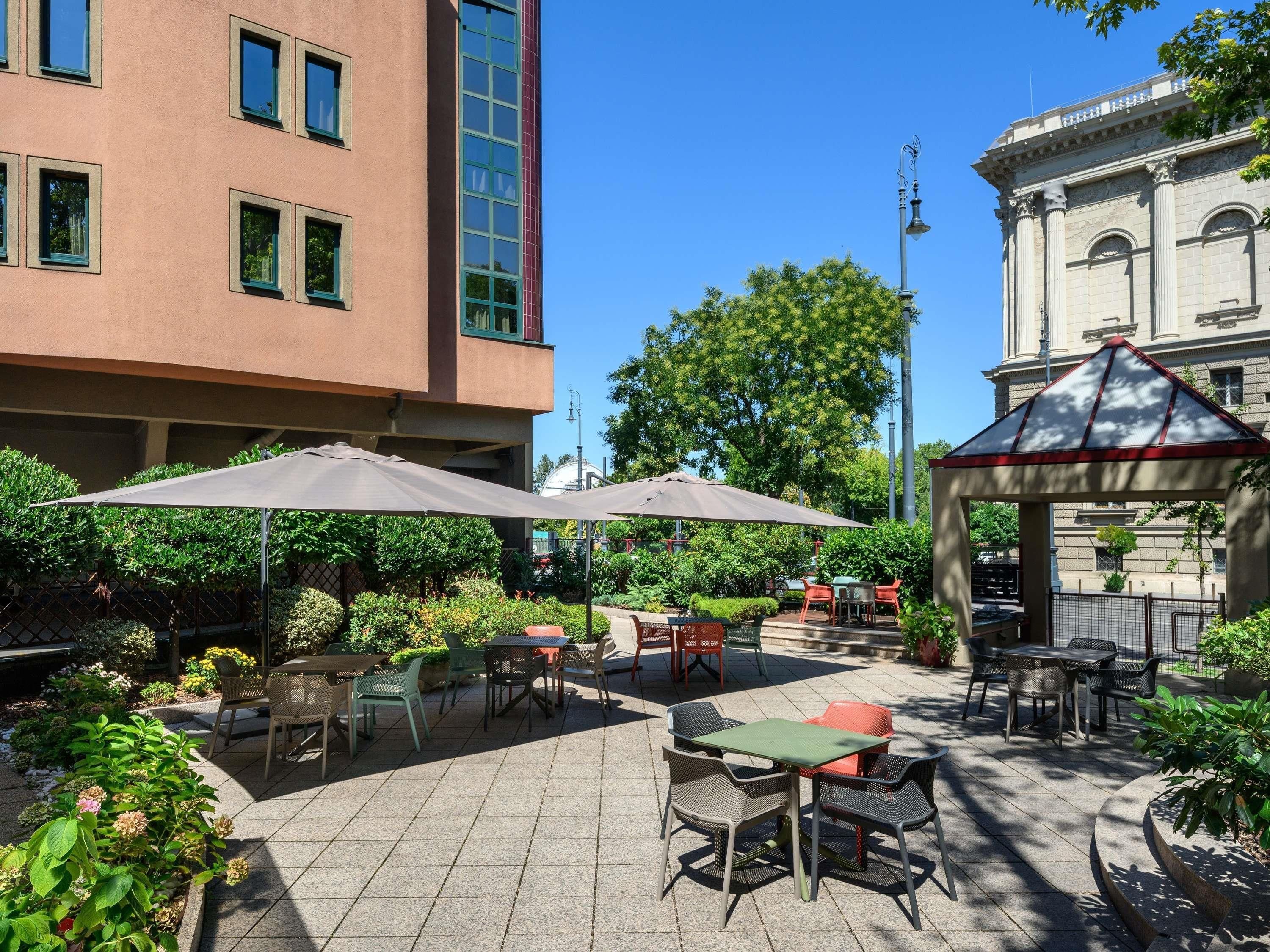 An outdoor patio area with tables and chairs, surrounded by greenery and a building with classical architecture.
