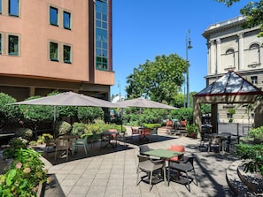 An outdoor patio area with tables and chairs, surrounded by greenery and a building with classical architecture.