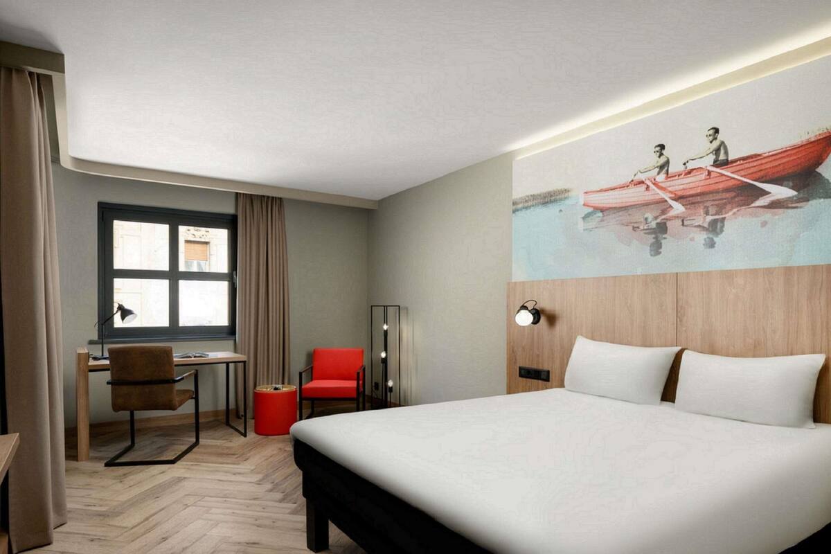 Hotellihuone, jossa on suuri sänky, työpöytä tuolilla, punainen tuoli, lamppu ja maalaus, jossa on kuvattuna soutajia veneessä.