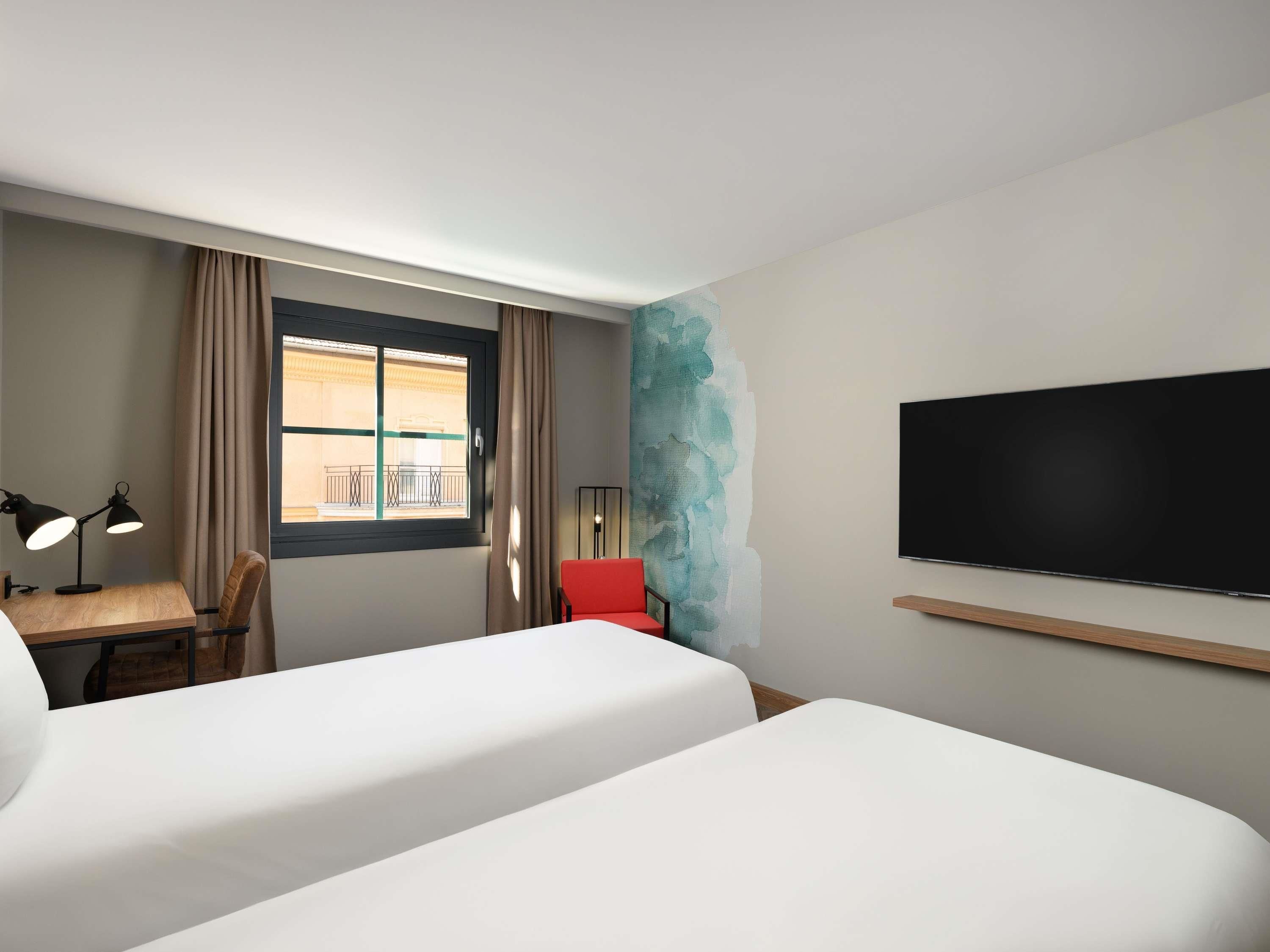 Una habitación de hotel con dos camas, un escritorio con lámpara, una silla roja, un televisor y una ventana con cortinas.