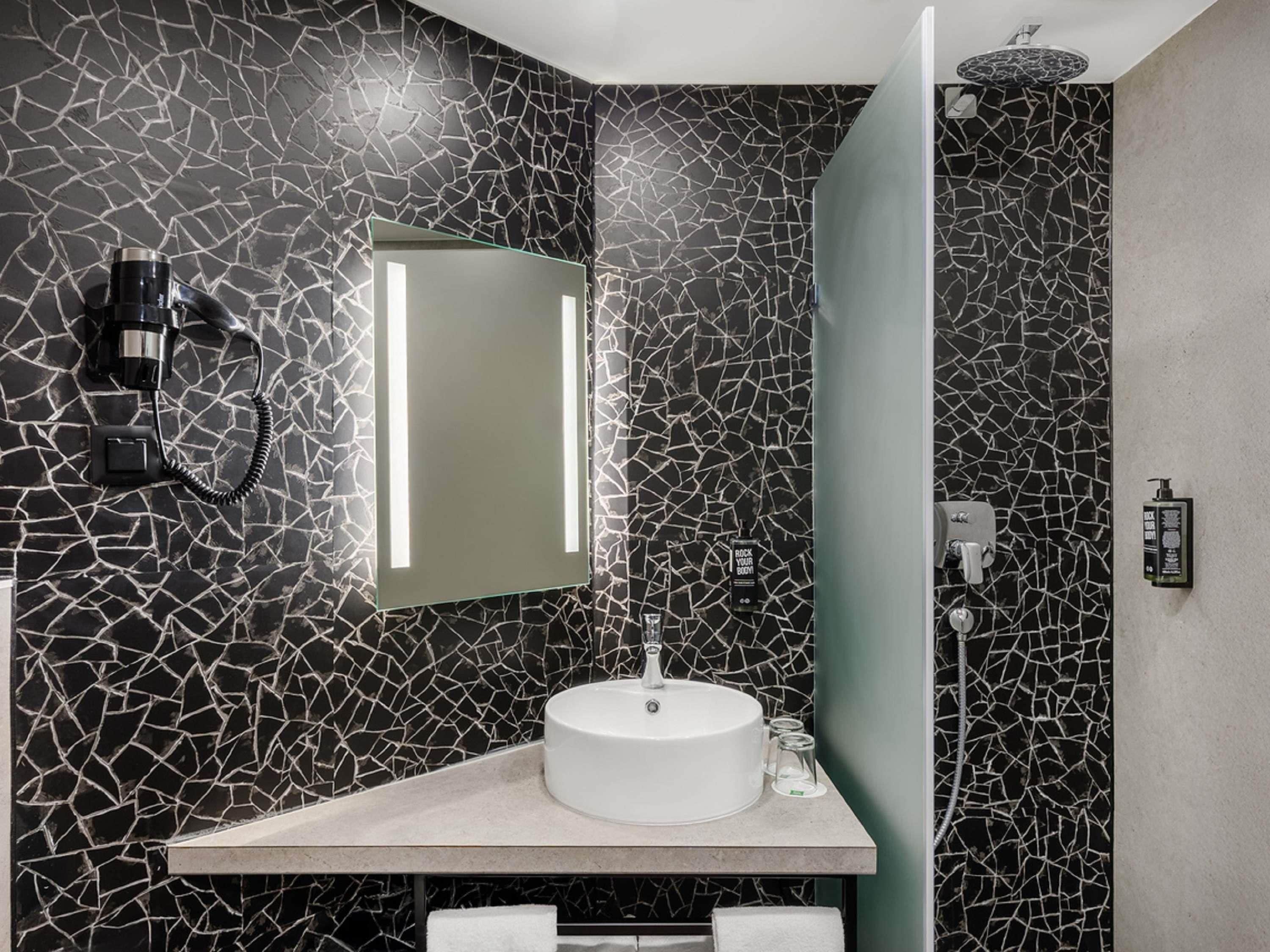 A bathroom with a cracked black wall, a white sink, a mirror, a hairdryer, and a shower.