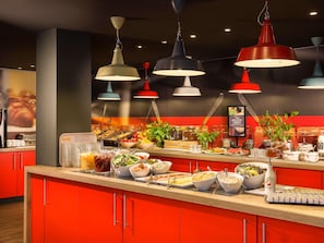 A buffet counter with various food items, a coffee machine, and hanging pendant lights. - ibis Styles Budapest Heroes Square (Budapest)