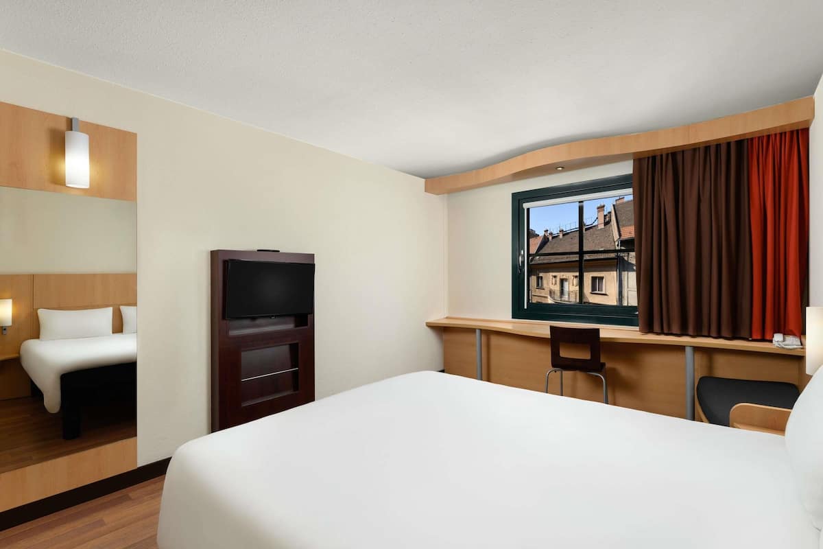 A hotel room with a bed, a TV, a desk, and a window with curtains.