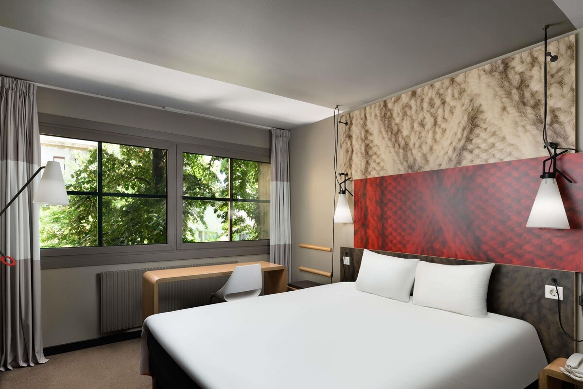 A hotel room with a bed, desk, and large windows.
